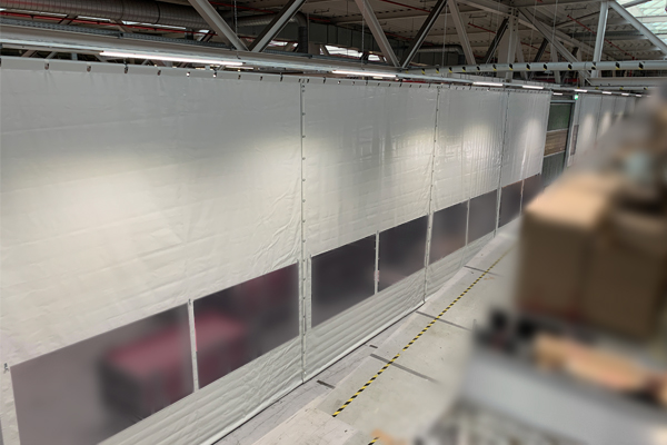 Am Stahlträger unter einem Dach einer Industriehalle befestigte Hallenabtrennung. Breite 25 m x Höhe 6 m aus grauer PVC-Plane mit Sichtfenstern im unteren Bereich.