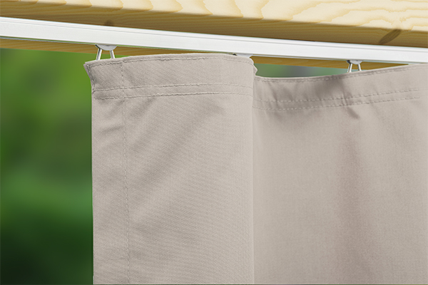 Wohnliches Flair: Der blickdichte Outdoor-Vorhang Gentle für gemütliches Terrassen-Ambiente. Detailaufnahme der Montage eines beigen Outdoor-Vorhangs Gentle an einer an einem Holzbalken von unten angeschraubten Vorhangschiene mit sanft fallendm Vorhangstoff.