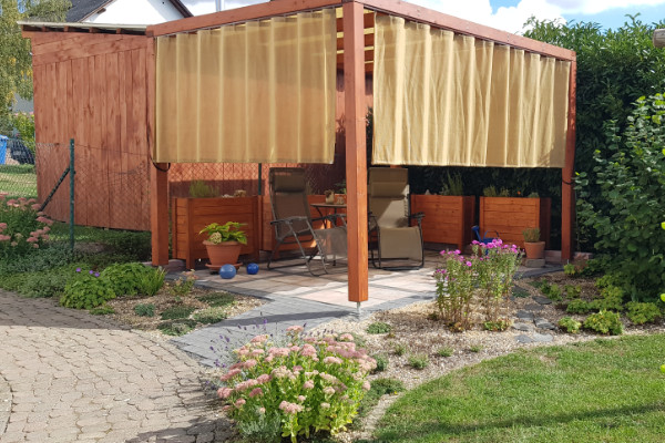 Holzpergola mit komplett geschlossenen, gelb-beigen Seitenvorhängen auf halber Höhe zum Schutz vor Schrägsonne