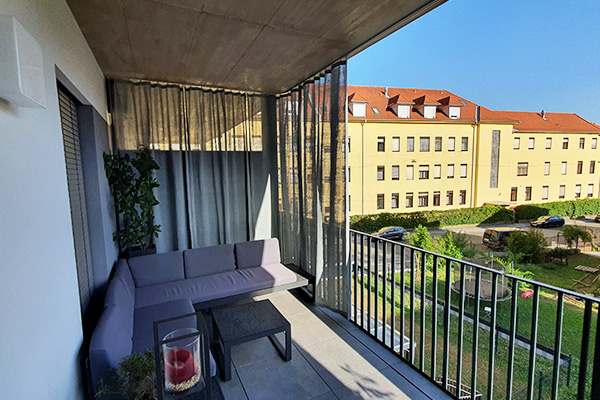 Der Aussenvorhang auf Balkonen schützt vor schräg stehender Sonne und Regen. Verschiebbarer Aussenvorhang in grau auf einem Balkon mit Lounge-Möbeln. Im Hintergrund ein Mehrfamilienhaus.