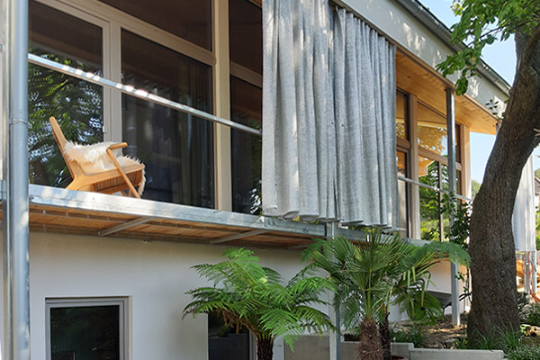 Fassaden-Design: Coolish Hitzeschutz-Vorhang für großflächige Verschattung von Glasflächen auf Balkonen und Terrassen. Außenansicht einer modernen Glasfassade hinter einem Balkon mit einem großflächigen Coolish Outdoor-Vorhang als Sonnenschutz.
